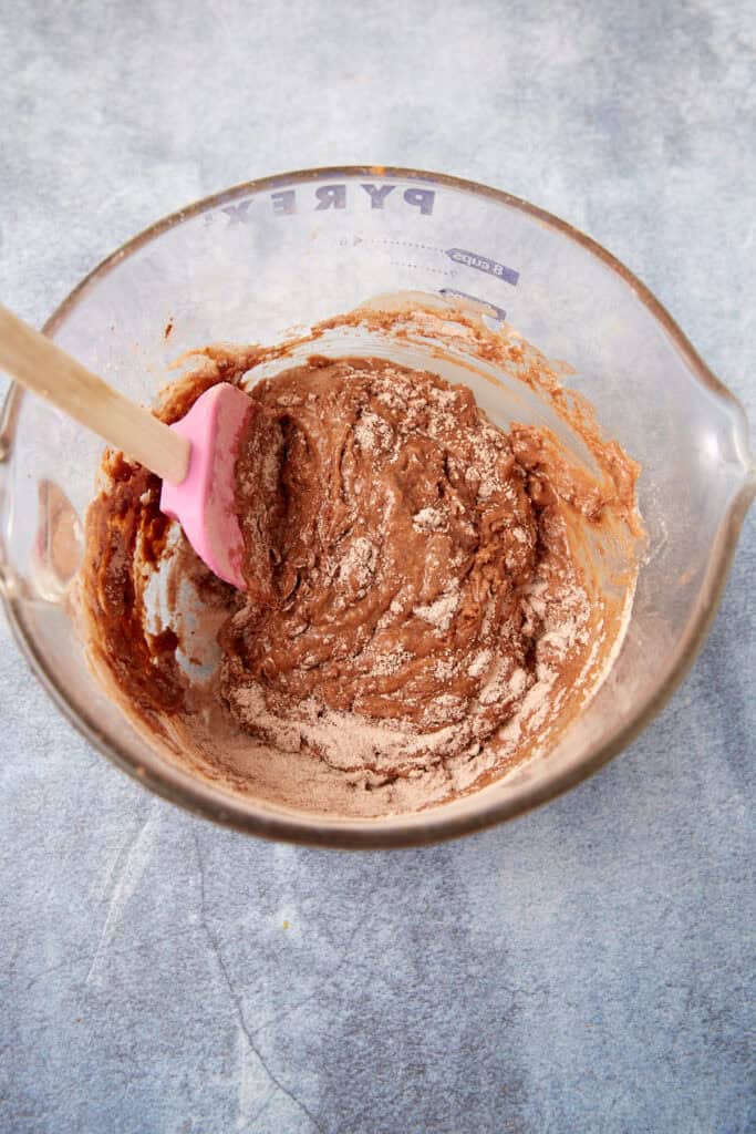 Mixing bowl of thick chocolate cake batter being stirred with a pink spatula, streaked with dry ingredients.