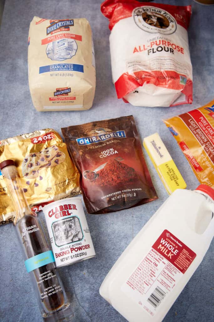Overhead view of slow cooker chocolate lava cake ingredients laid out on a blue surface, including sugar, flour, cocoa powder, chocolate chips, baking powder, brown sugar, butter, milk, and vanilla.