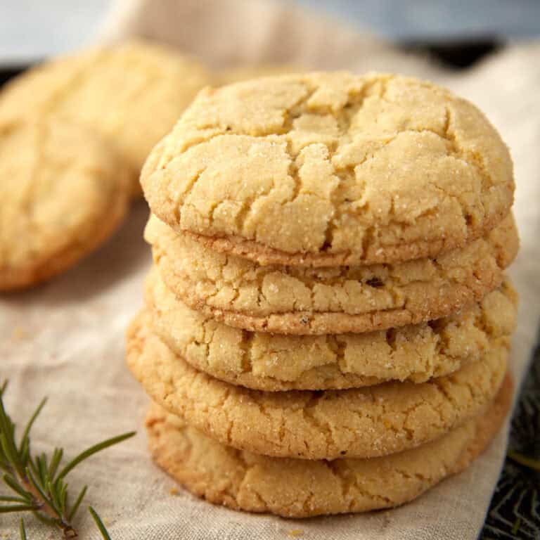 Stack of golden lemon cornmeal cookies with crackly tops on a linen napkin, with a rosemary sprig beside them.