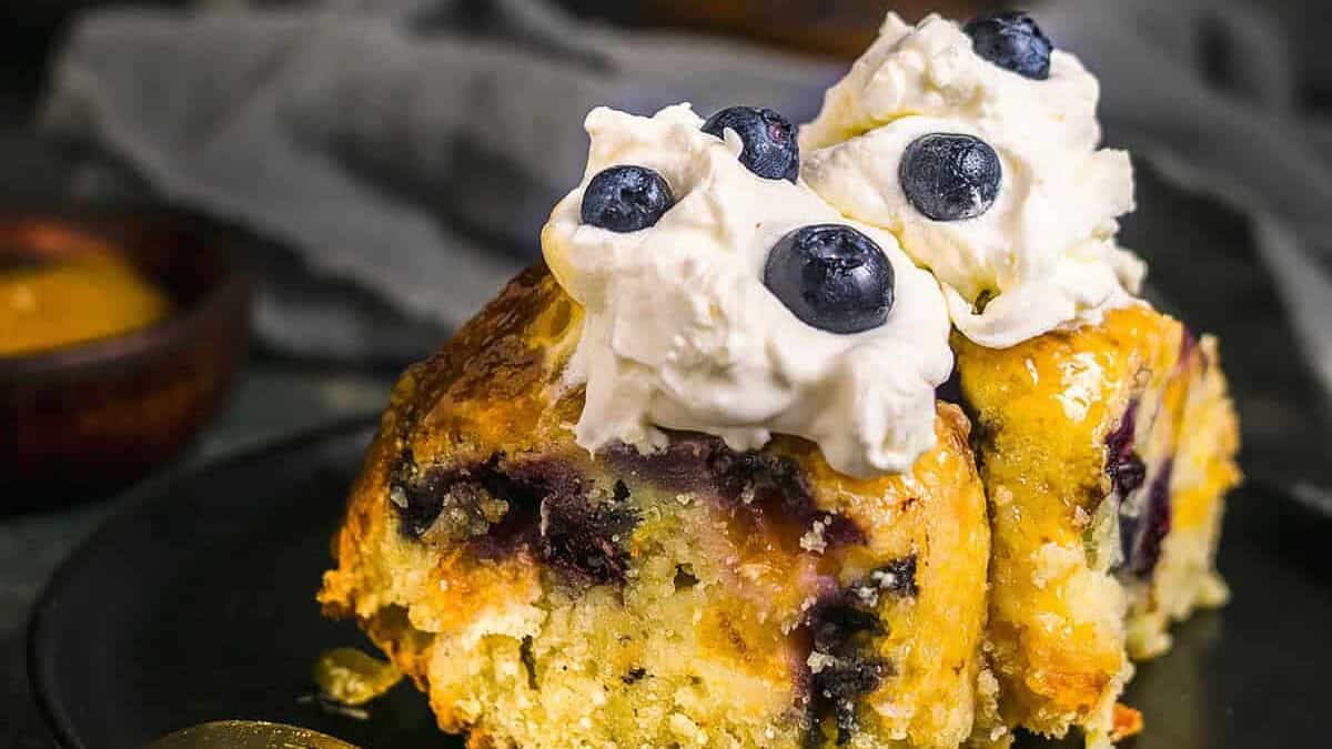 Close-up of a slice of peach blueberry cake topped with whipped cream and fresh blueberries on a black plate.