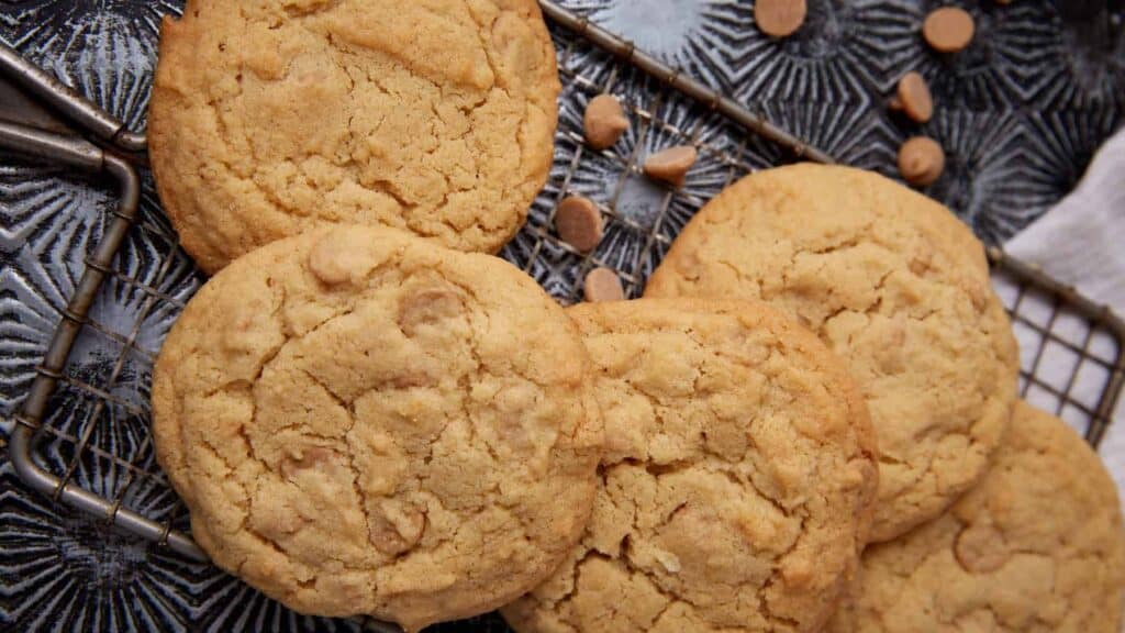 Stack of Reese&rsquo;s peanut butter cookies cooling on a small wire rack over a dark patterned tray, with peanut butter chips sprinkled nearby.