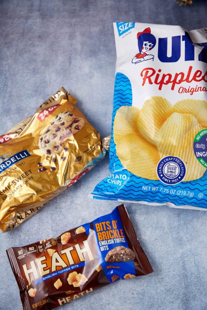 Bags of semi-sweet chocolate chips, rippled potato chips, and Heath English toffee bits on a blue surface for toffee chocolate chip cookies.