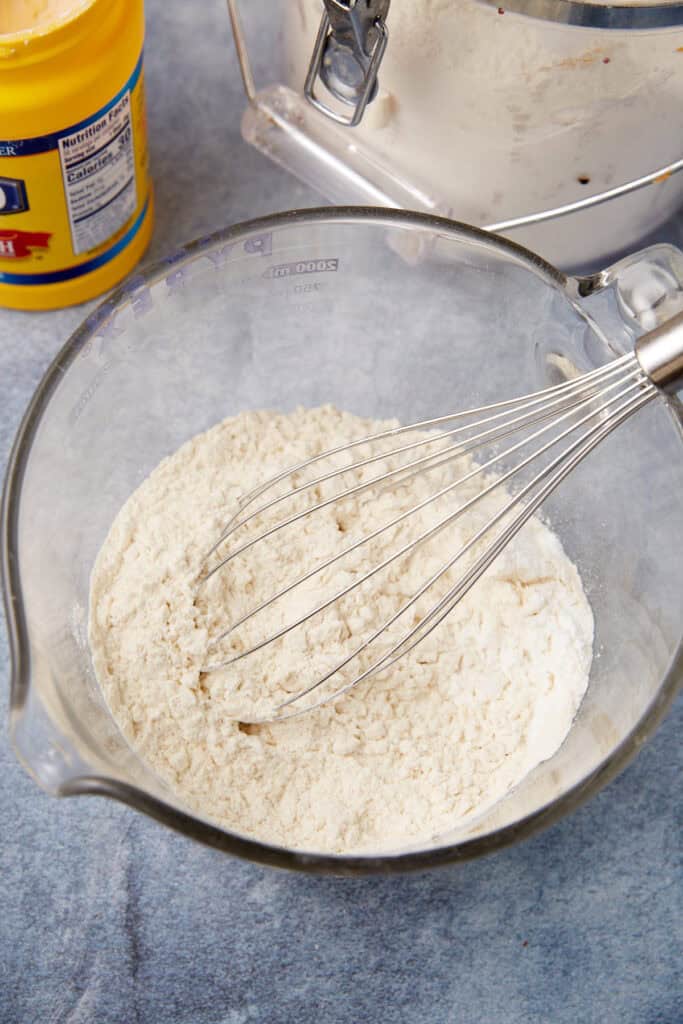 Glass bowl of flour mixture being whisked for toffee chocolate chip cookies, with cornstarch container and flour canister nearby.