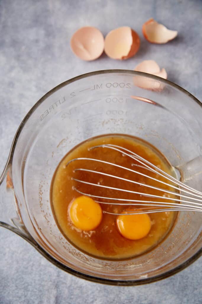 Glass mixing bowl with melted butter and sugar mixture and two egg yolks ready to be whisked together, eggshells in the background.