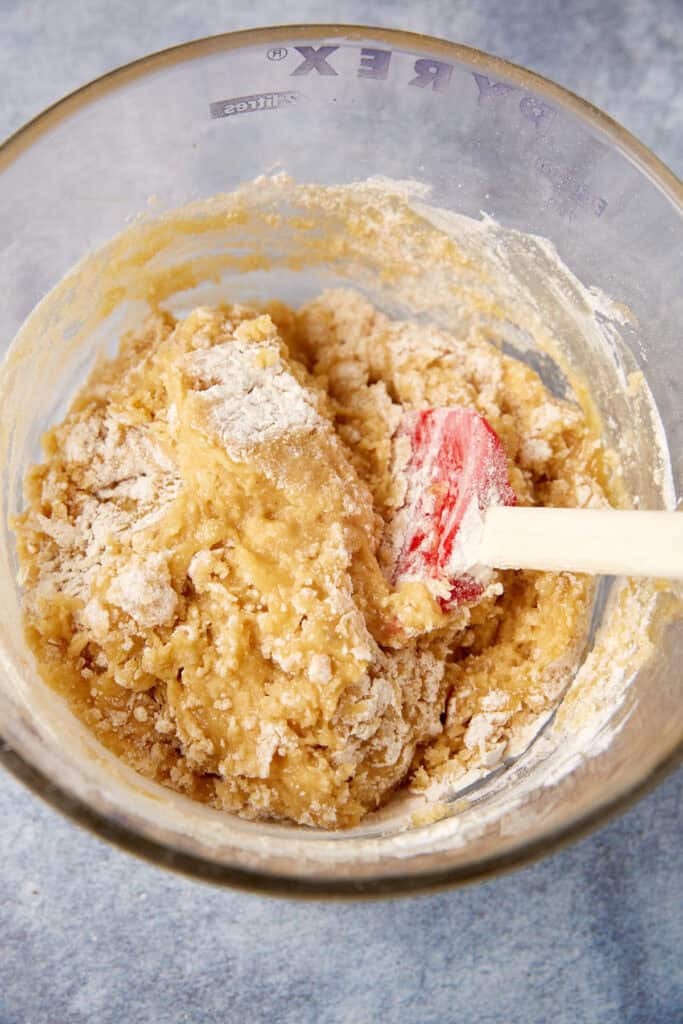 Cookie dough in a glass bowl with flour still visible on top as it is being folded together with a red spatula.