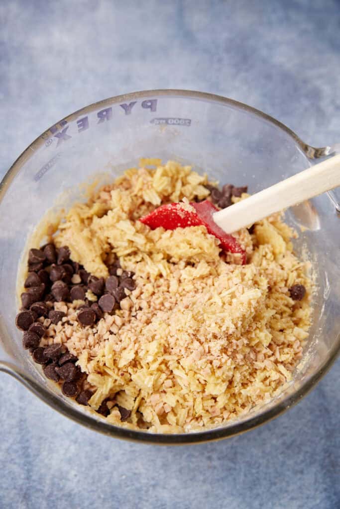 Glass mixing bowl filled with cookie dough, crushed potato chips, chocolate chips, and toffee bits being folded together with a red spatula.