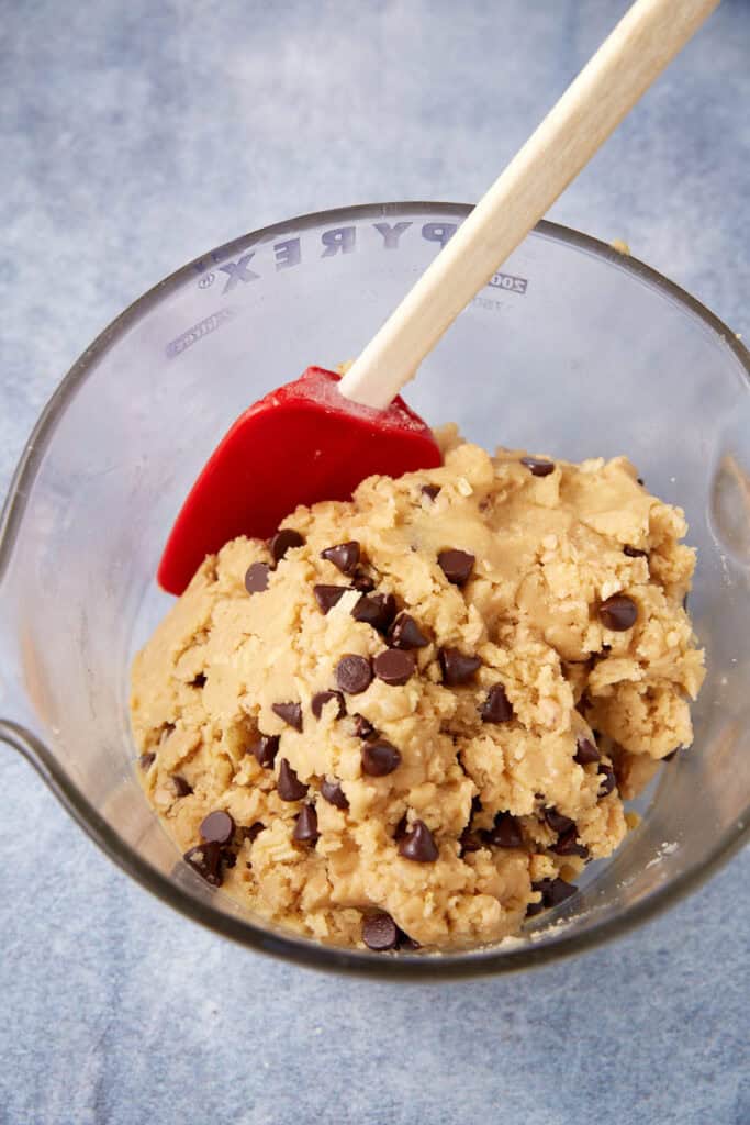 Thick toffee chocolate chip cookie dough in a glass bowl with a red spatula, chocolate chips evenly mixed throughout.