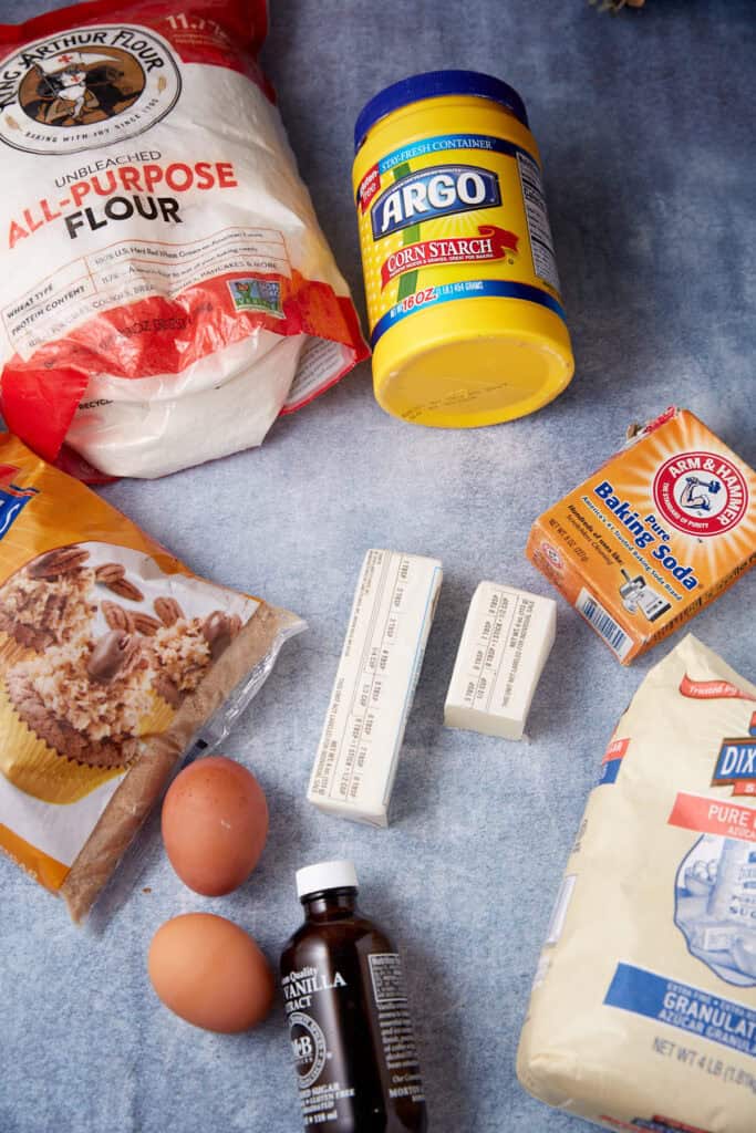 Flour, cornstarch, sugar, brown sugar, butter sticks, baking soda, eggs, and vanilla extract arranged on a blue surface for toffee chocolate chip cookies.