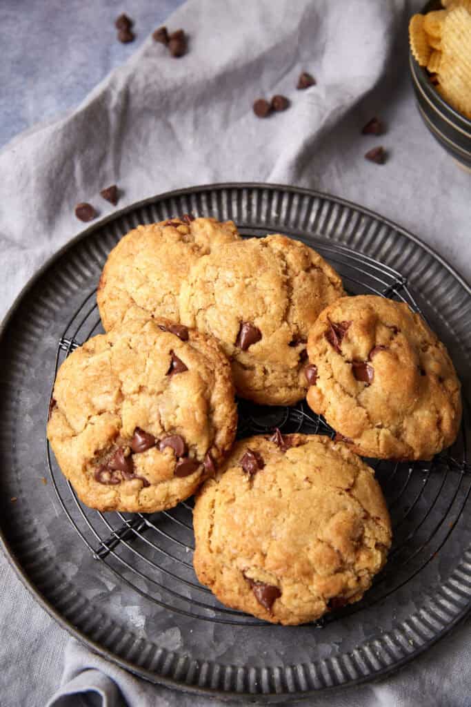 Toffee chocolate chip cookies on a round cooling rack with a gray linen and scattered chocolate chips in the background.
