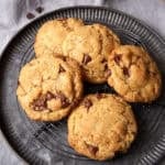 Pile of toffee chocolate chip cookies cooling on a round wire rack set inside a metal tray.