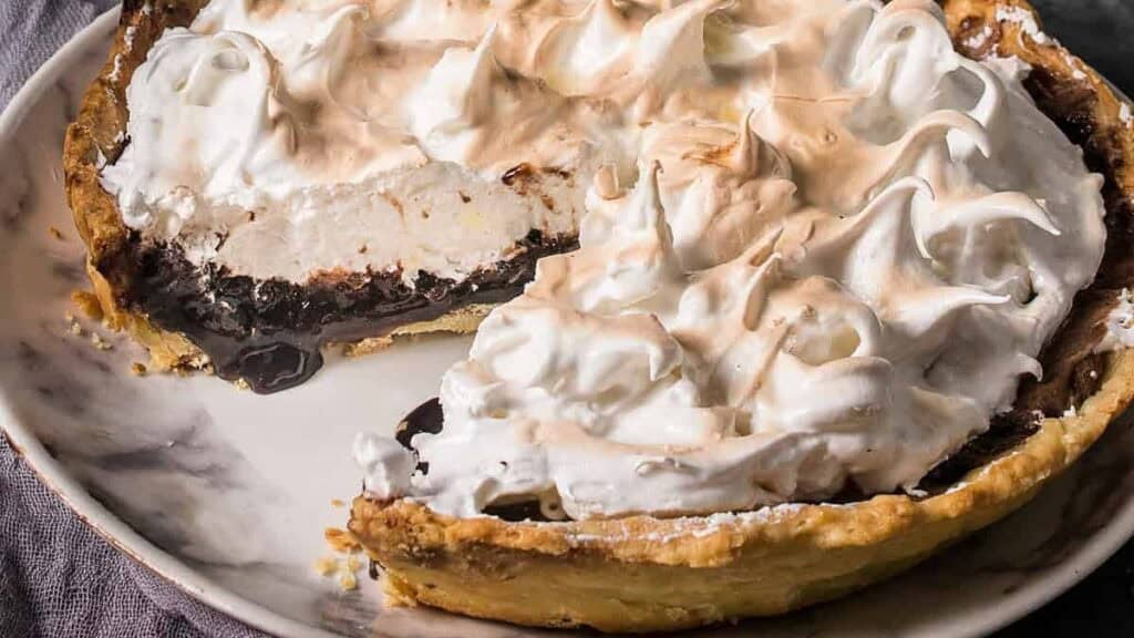 Whole chocolate meringue pie with a slice taken out, set on a plate with a cloth napkin beside it.