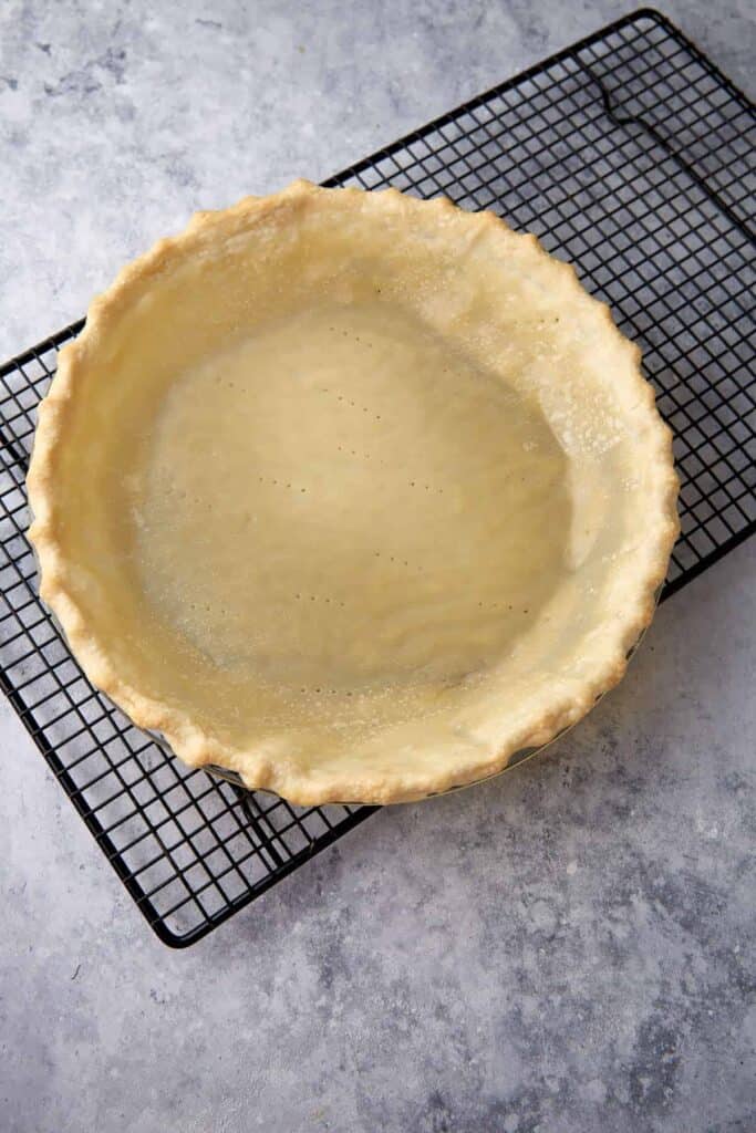 A partially baked pie crust with fork holes cools on a wire rack.