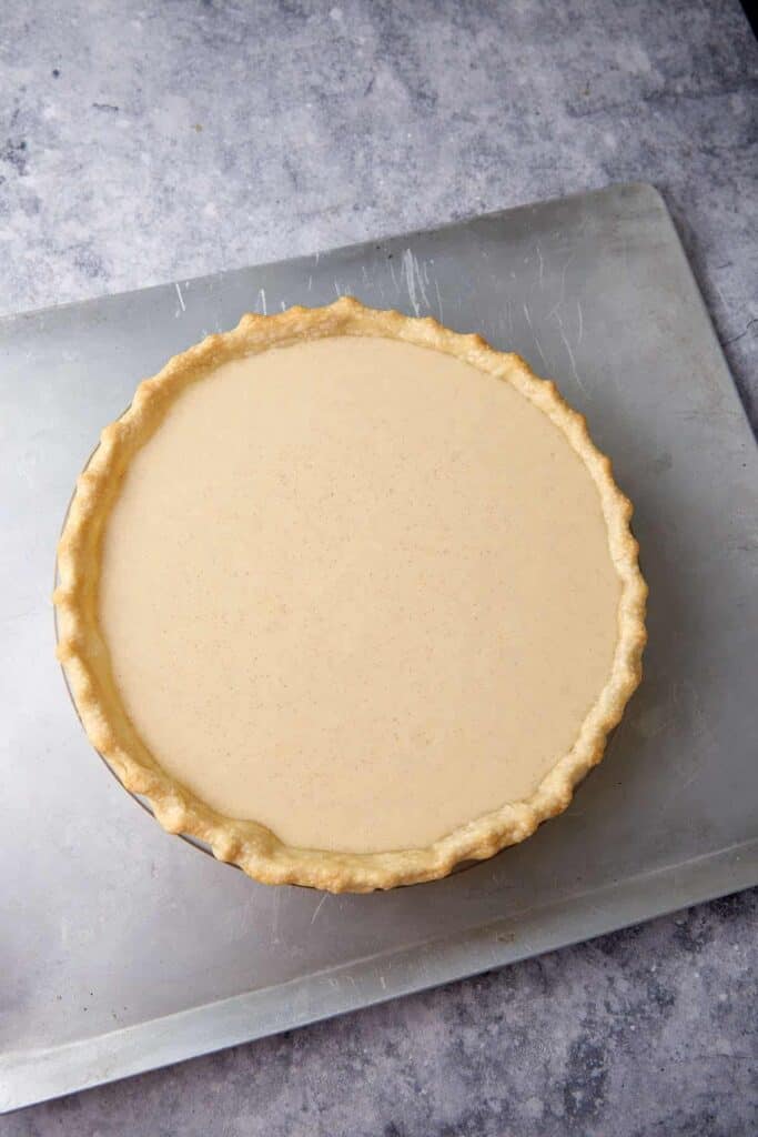 Unbaked buttermilk pie filling poured into a pie crust in a glass pie plate sitting on a metal baking sheet.