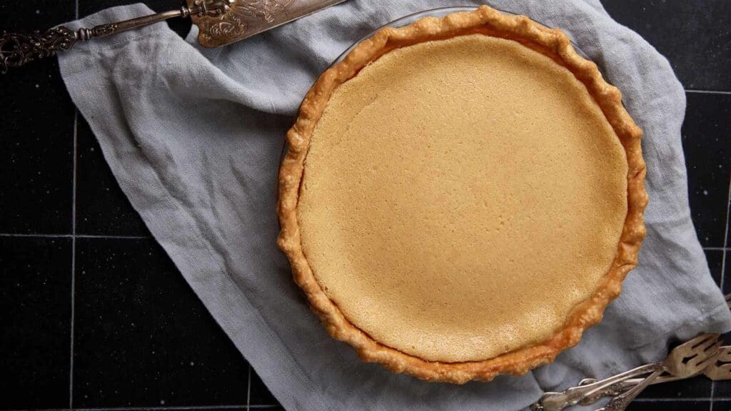 Whole baked buttermilk pie with a smooth custard top on a gray linen cloth beside a pie server.