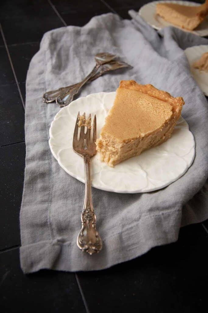 A slice of buttermilk pie sits on a white plate with a fork on a gray cloth.