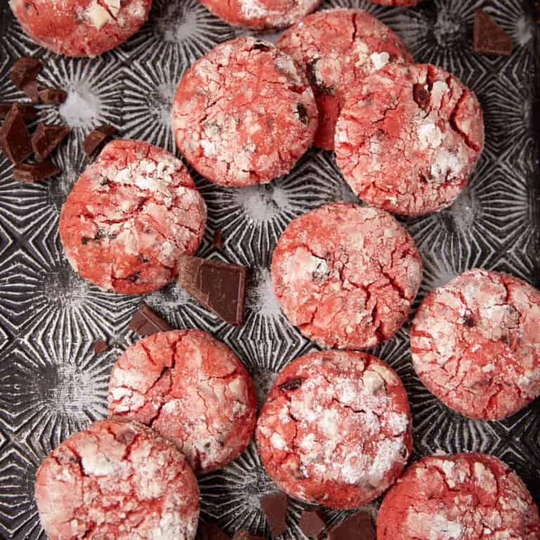 Pink cherry chocolate crinkle cookies scattered on a patterned tray with chunks of dark chocolate around them.