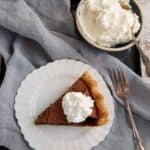 A slice of chocolate chess pie on a white plate is topped with whipped cream beside a bowl of whipped cream.