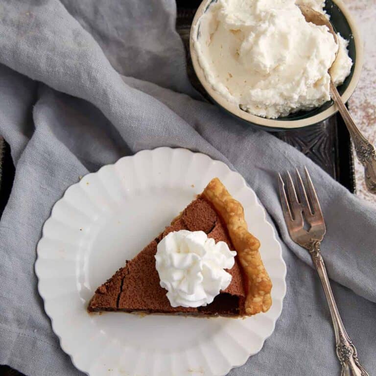 A slice of chocolate chess pie on a white plate is topped with whipped cream beside a bowl of whipped cream.