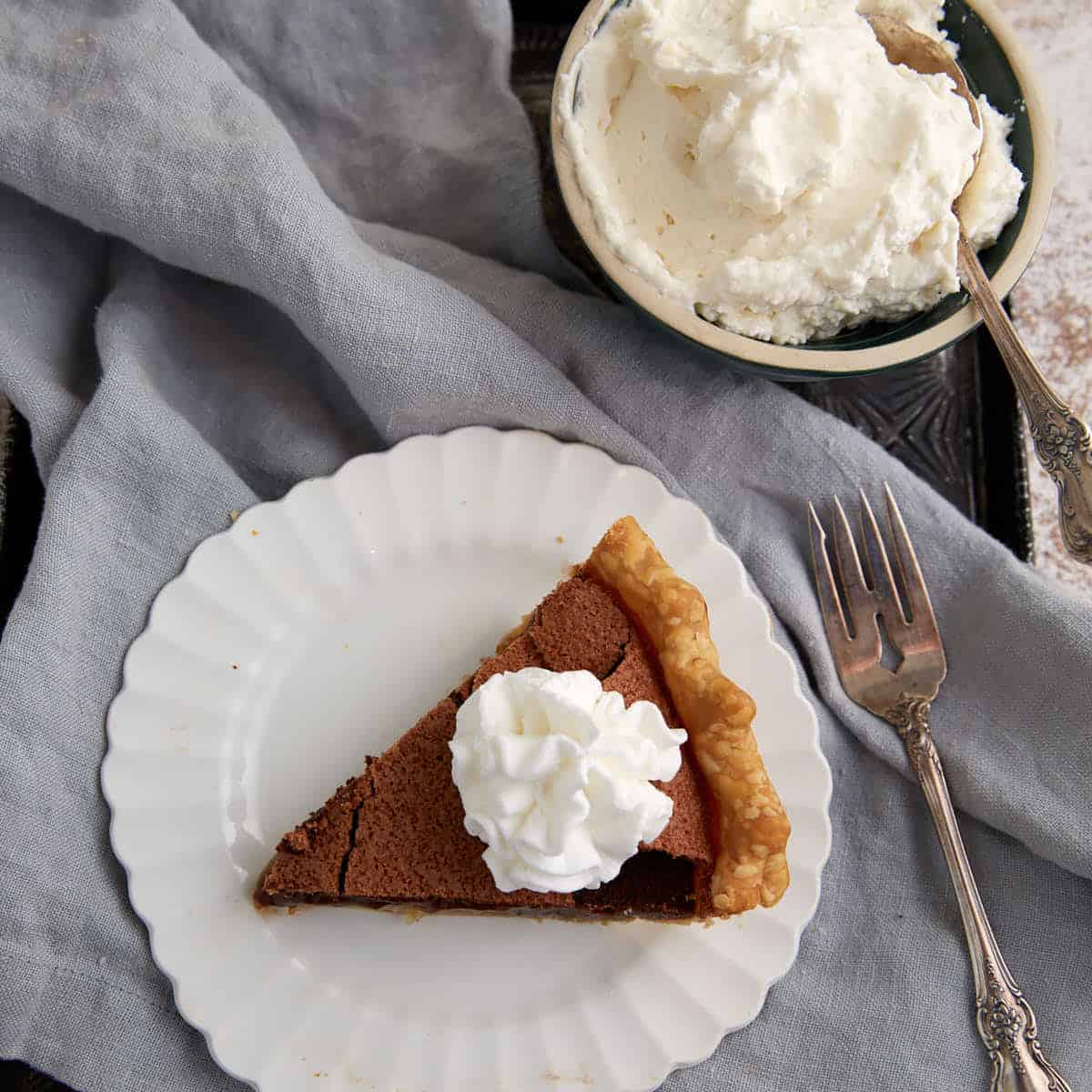 A slice of chocolate chess pie on a white plate is topped with whipped cream beside a bowl of whipped cream.