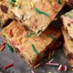 Stack of sprinkle blondie squares on a dark plate with a few loose rainbow sprinkles around them.
