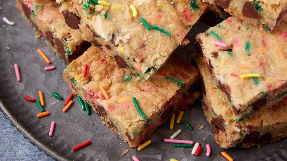 Stack of sprinkle blondie squares on a dark plate with a few loose rainbow sprinkles around them.