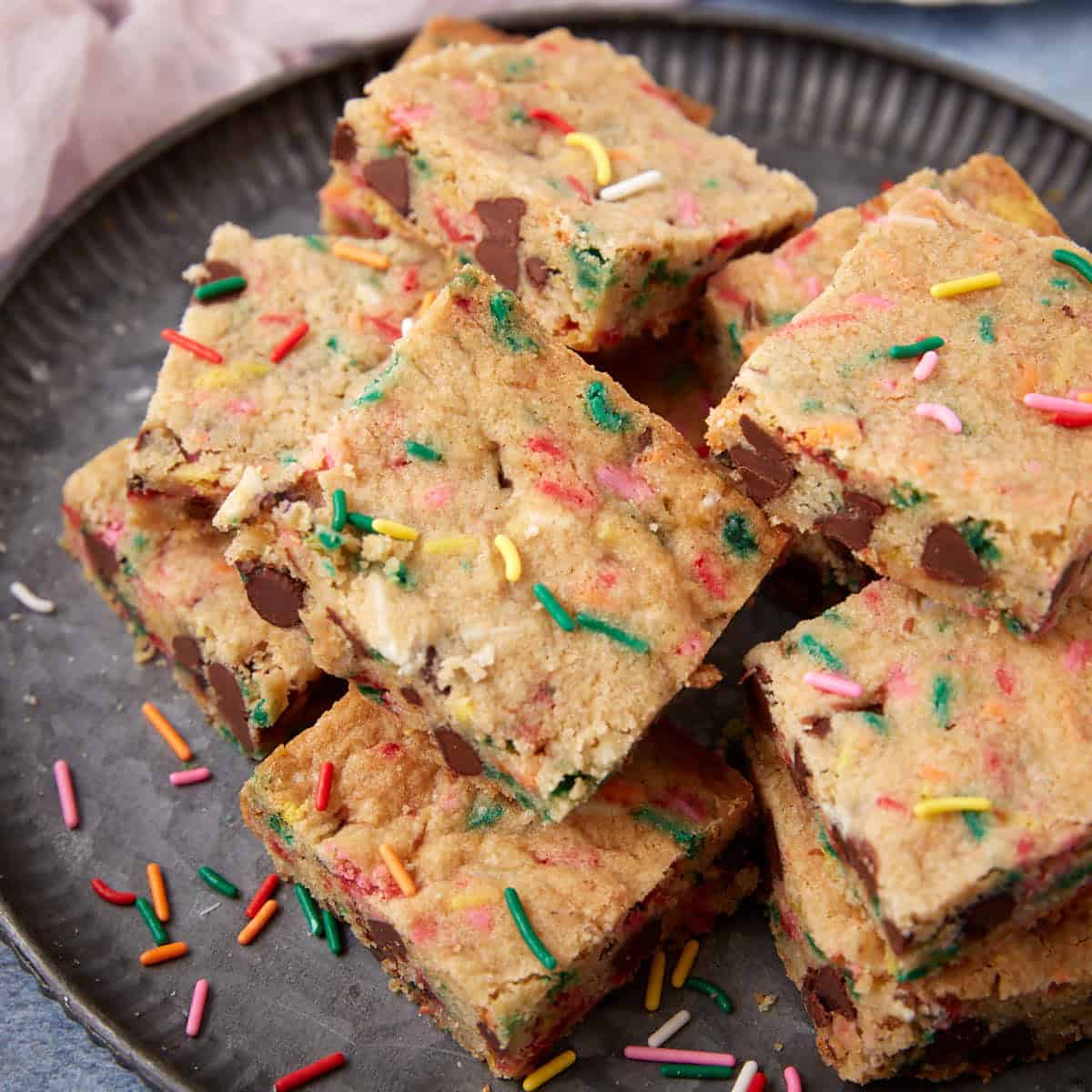 Several sprinkle blondie squares piled on a plate with scattered rainbow sprinkles nearby.