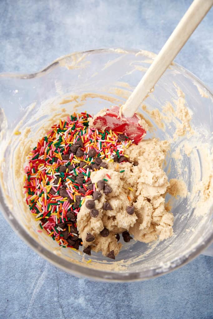 Mixing bowl with blondie dough and a pile of rainbow sprinkles and chocolate chips, ready to be folded in.