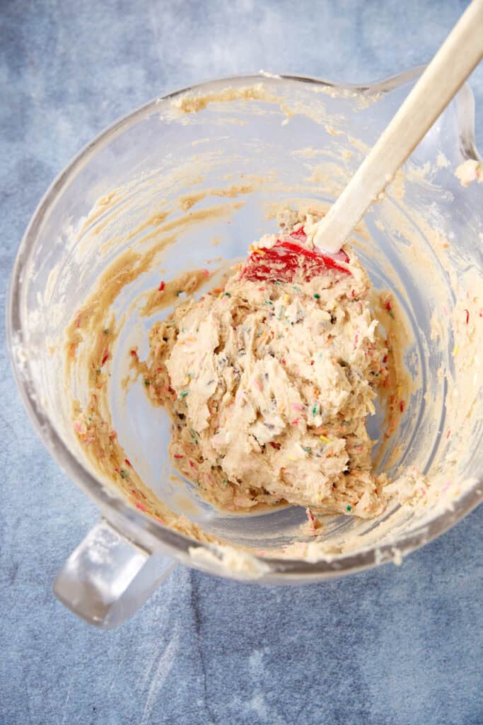 Cookie dough for sprinkle blondies in a mixing bowl with a spatula, showing chocolate chips and sprinkles mixed throughout.