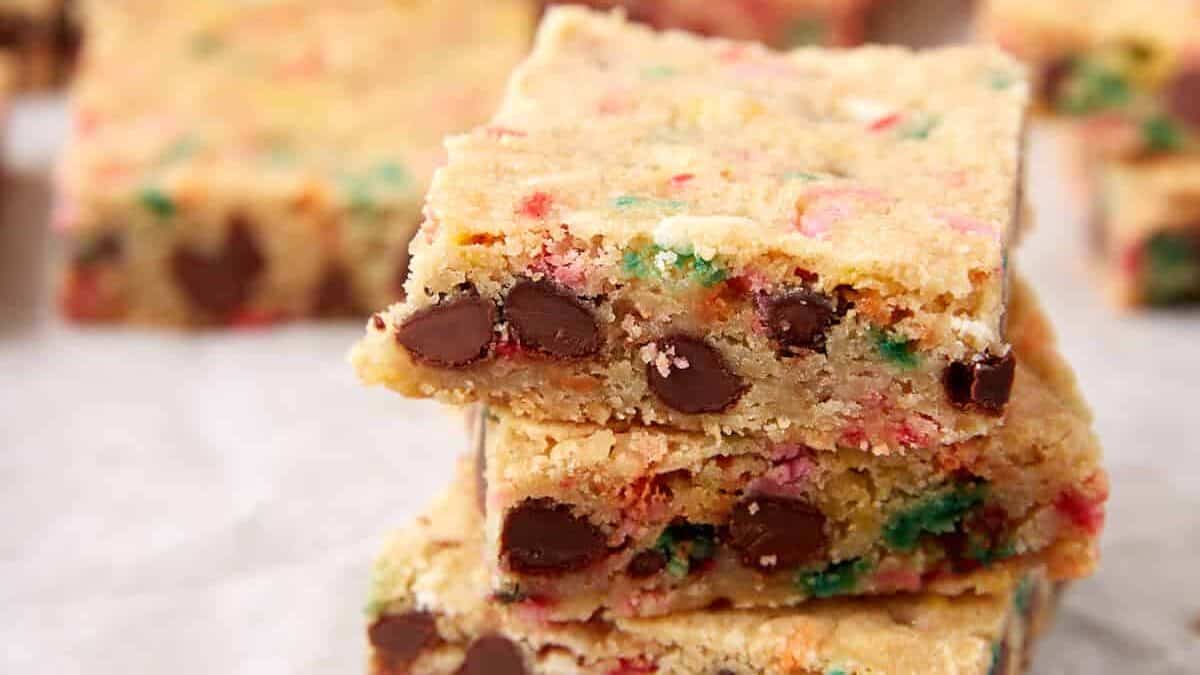 Three sprinkle blondie squares stacked on parchment paper, with more blondies blurred in the background.