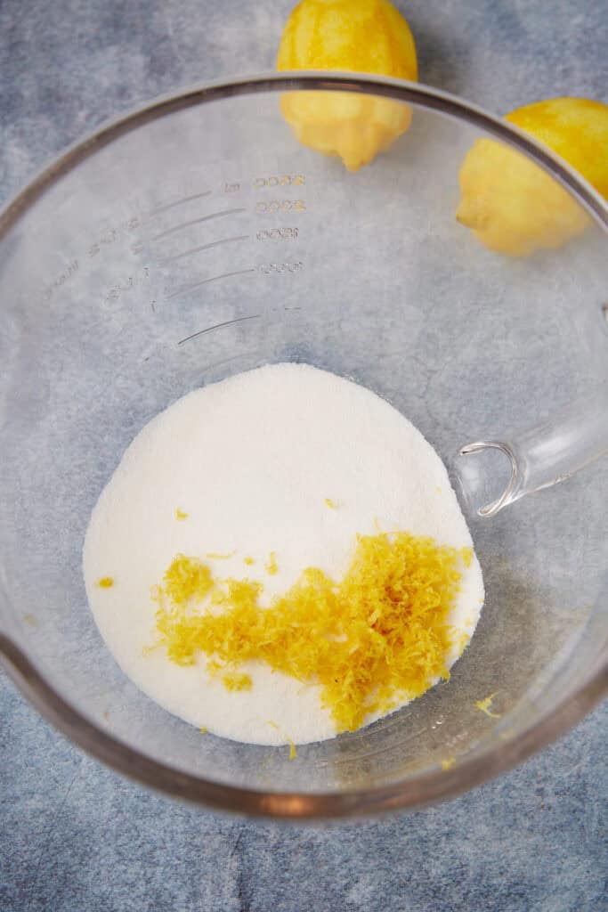 Granulated sugar and fresh lemon zest in a mixing bowl with whole lemons in the background.