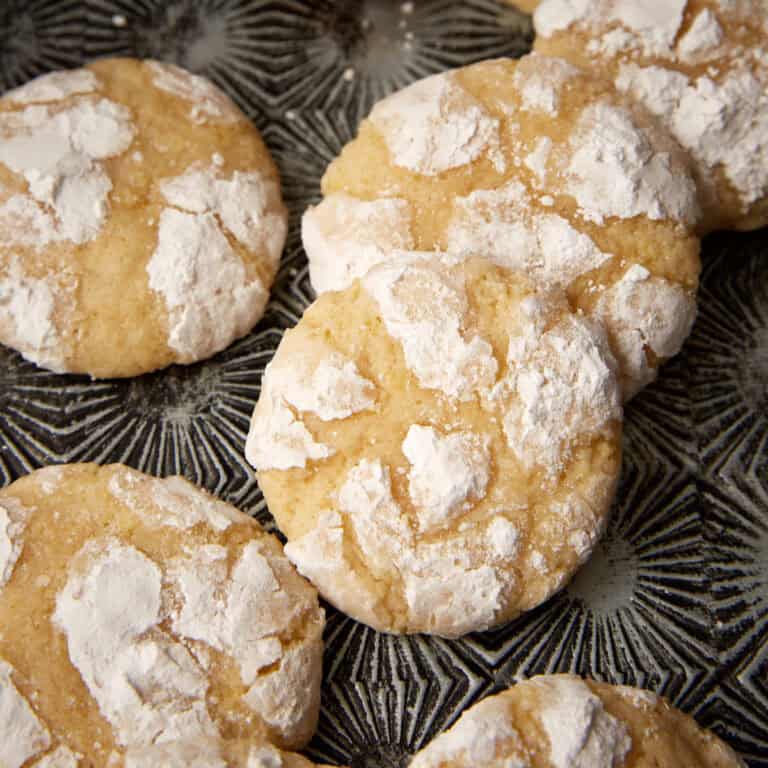 Close-up of lemon crinkle cookies with thick powdered sugar cracks on a dark patterned plate.