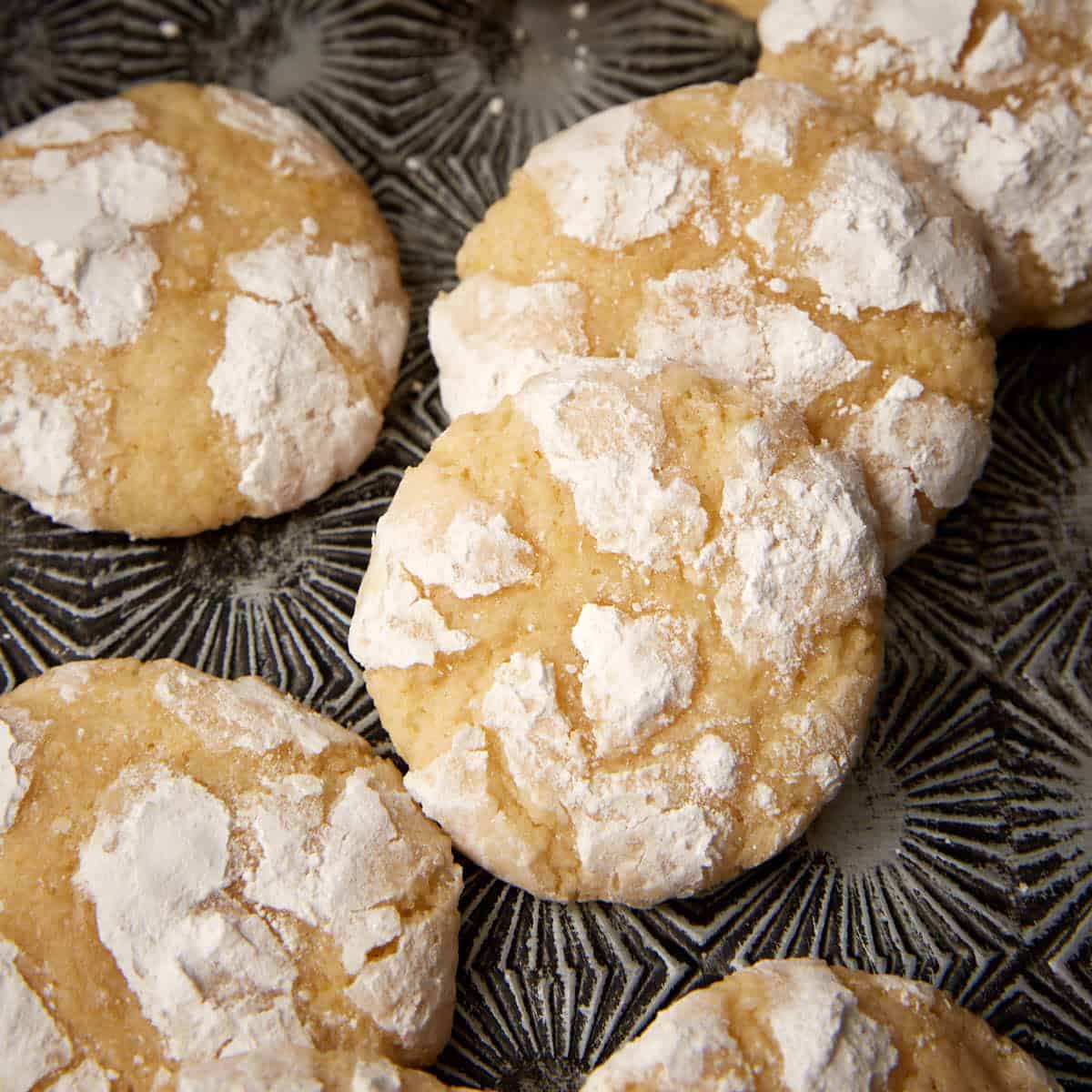 Close-up of lemon crinkle cookies with thick powdered sugar cracks on a dark patterned plate.