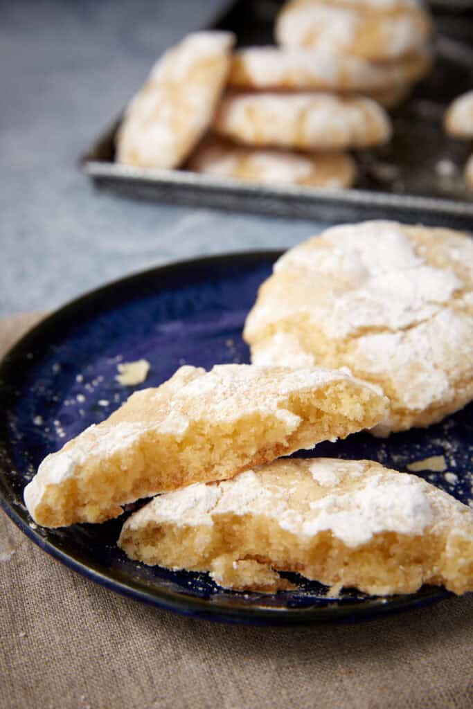 Broken lemon crinkle cookie on a dark blue plate showing the soft, chewy center; baking tray blurred behind.