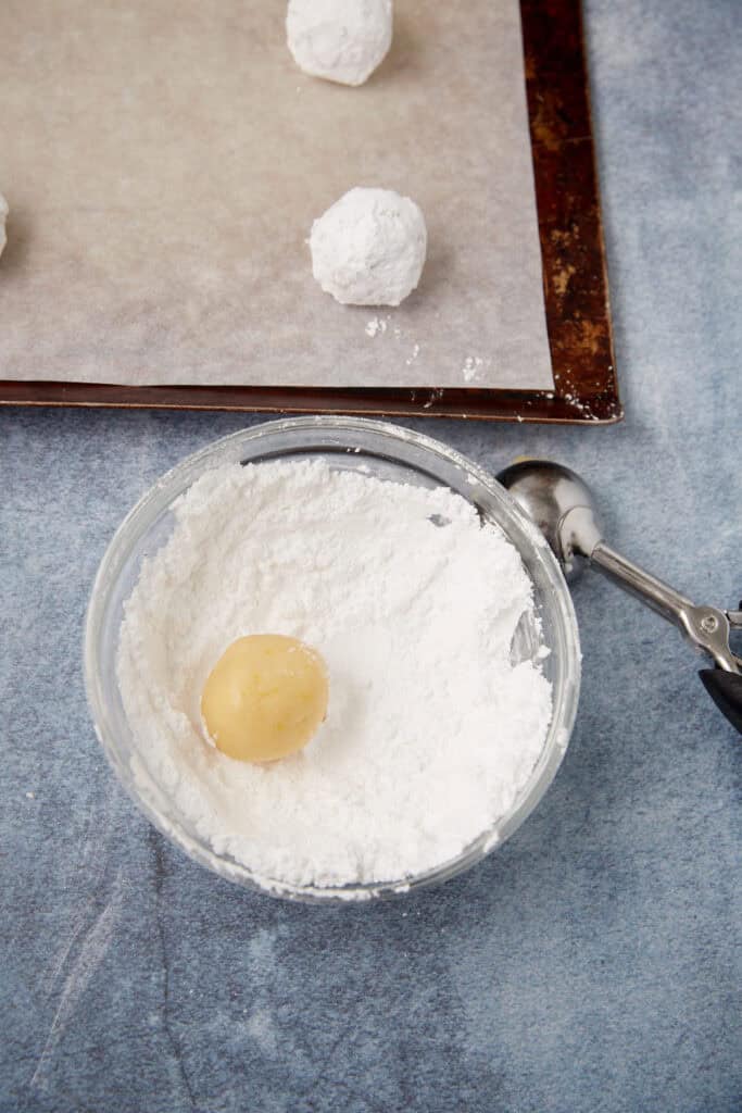 Small bowl of powdered sugar with a cookie dough ball and a cookie scoop beside a baking sheet.