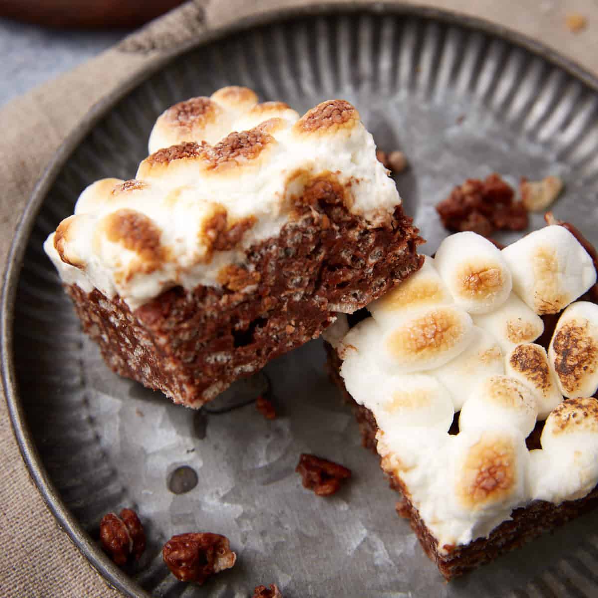 Two chocolate Rice Krispie s’mores bars topped with toasted mini marshmallows on a metal plate.