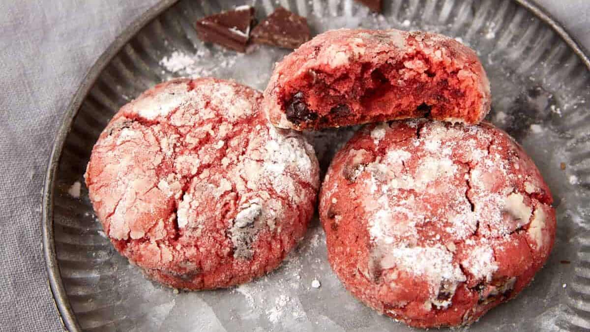 Three pink cherry chocolate crinkle cookies on a plate, with one cookie broken open to show the soft center.