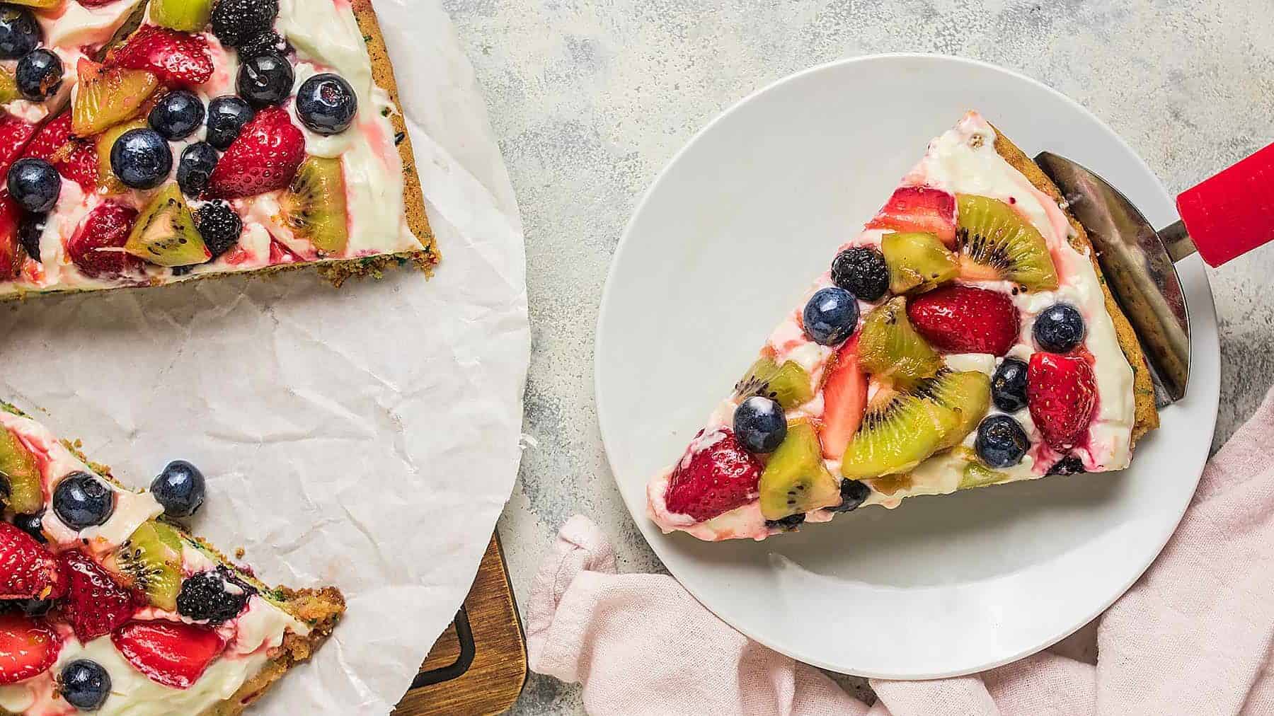 Single slice of fruit pizza served on a plate with a metal pie server and pink linen in the background.