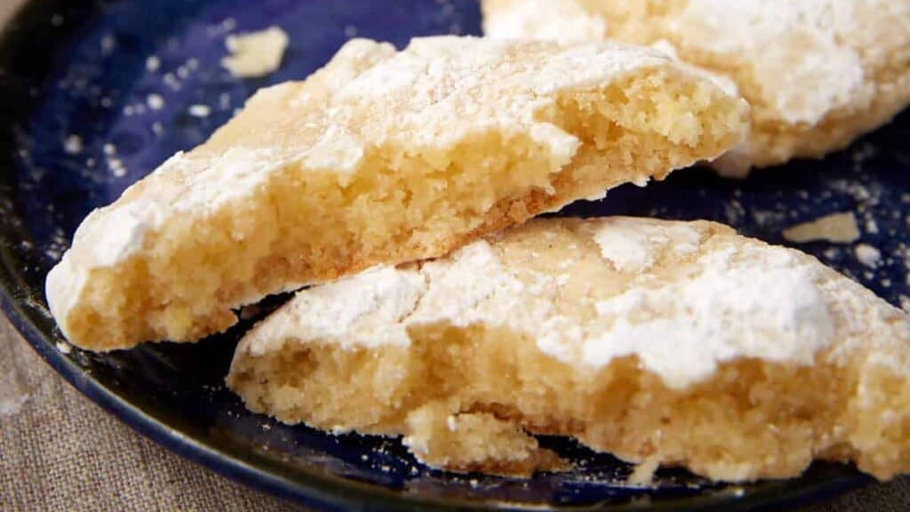 Broken lemon crinkle cookie on a dark blue plate showing the soft, chewy center; baking tray blurred behind.