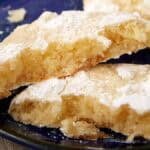 Broken lemon crinkle cookie on a dark blue plate showing the soft, chewy center; baking tray blurred behind.