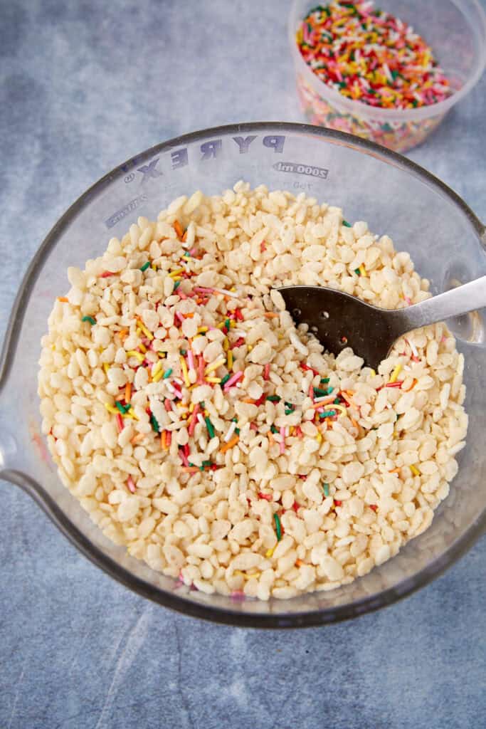 Rice Krispies cereal mixed with colorful sprinkles in a clear glass bowl with a spoon.