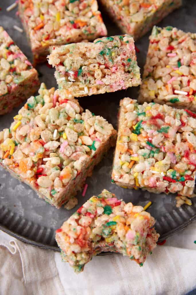 Squares of Rice Krispie treats with sprinkles served on a dark plate with one piece cut open.