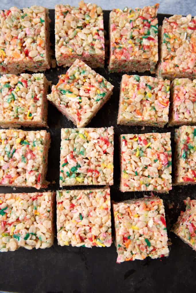Rice Krispie treats with sprinkles cut into squares and arranged on a dark tray.