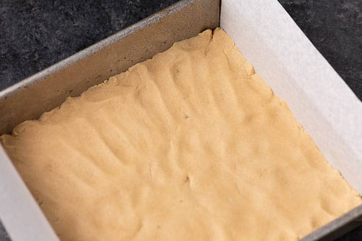 Shortbread crust pressed into a parchment-lined square baking pan before baking.