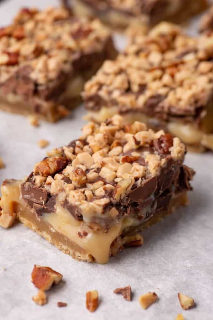 Close-up of a shortbread toffee bar with chocolate, chopped pecans, and toffee bits on parchment paper.
