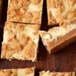 Carrot cake cheesecake bars cut into squares on a wooden board with one bar turned to show the layers.