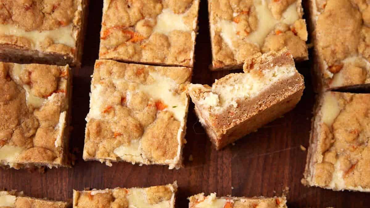 Carrot cake cheesecake bars cut into squares on a wooden board with one bar turned to show the layers.
