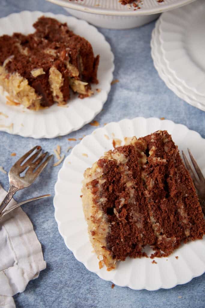 Slice of German chocolate cake on a white dessert plate with another plated slice and the whole cake in the background.