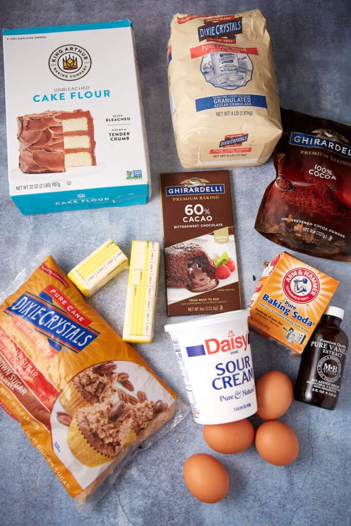 Ingredients for German chocolate cake arranged on a blue surface, including cake flour, cocoa powder, chocolate, sour cream, butter, sugar, eggs, and vanilla.