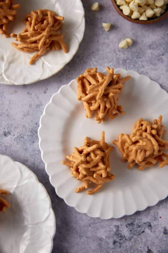 White chocolate haystack cookies arranged on white plates with white chocolate chips scattered nearby.
