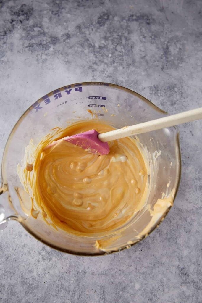 Melted white chocolate and butterscotch mixture stirred smooth in a glass measuring bowl for haystack cookies.